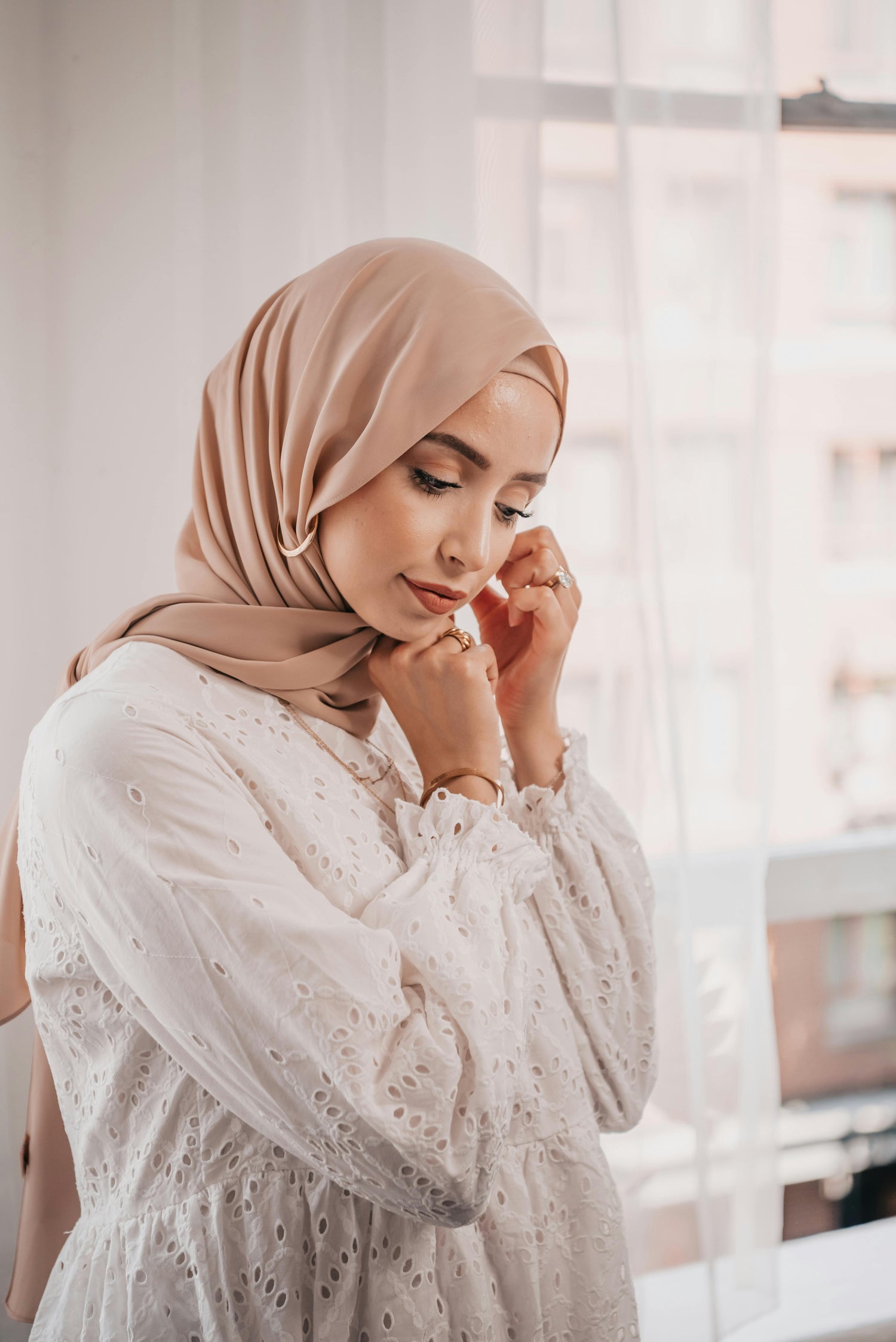 Woman adjusting taupe chiffon hijab in soft natural light, elegant modest fashion look with lightweight draped hijab
