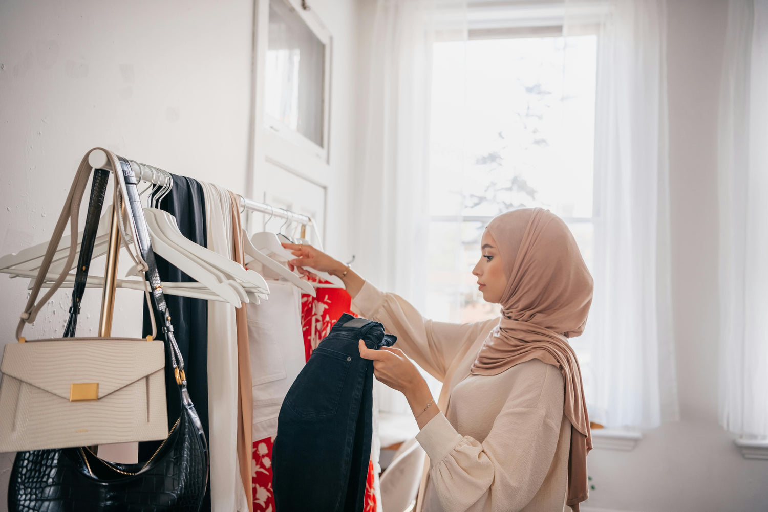 Woman wearing taupe hijab choosing outfit from wardrobe, modest fashion styling with neutral chiffon hijab and everyday wear