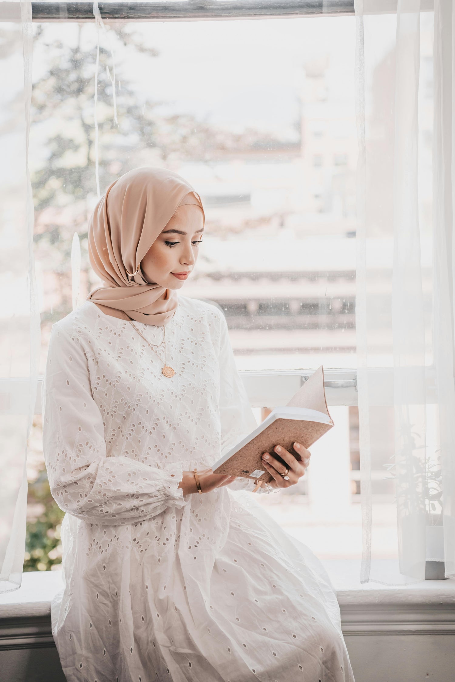 Woman wearing beige chiffon hijab reading by window in soft natural light, elegant modest fashion with lightweight draped hijab