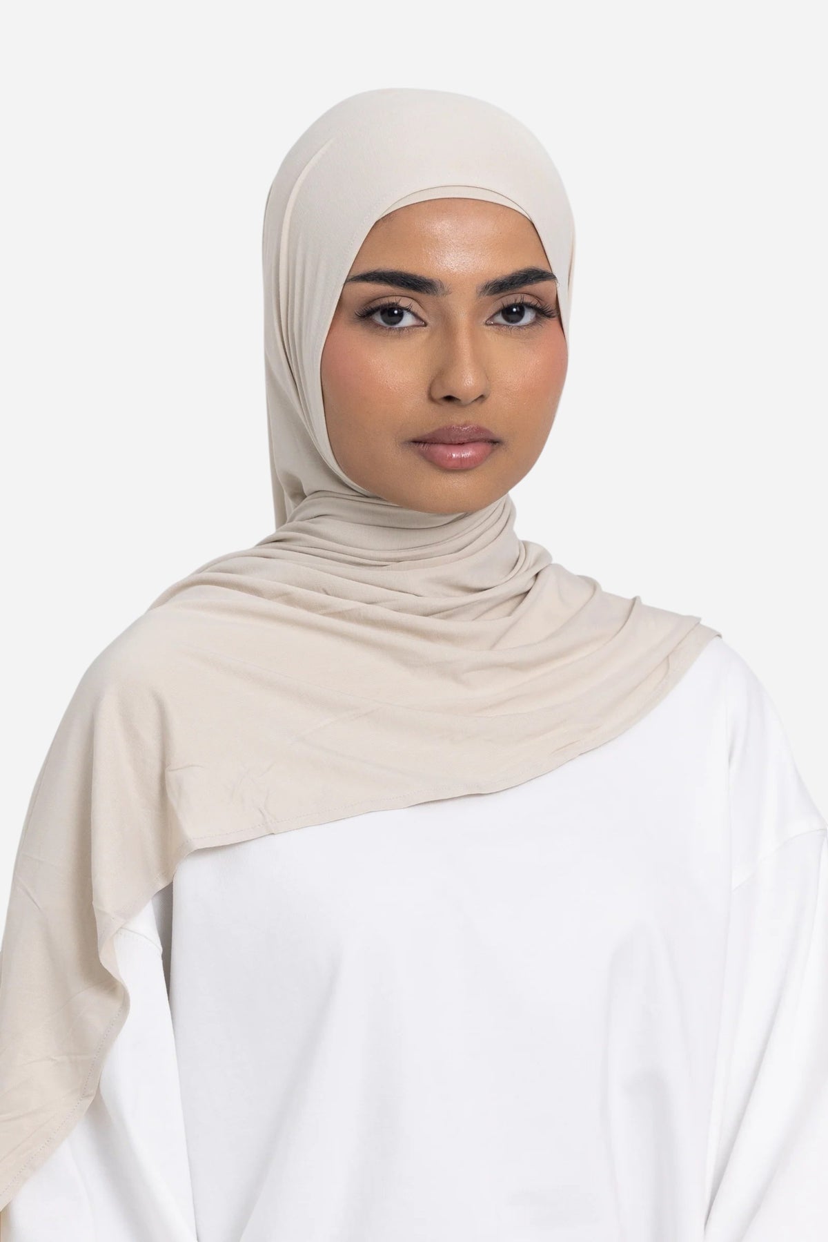 A woman wearing a light beige hijab and a white top, photographed against a plain light gray studio background.