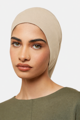 A close-up of a woman wearing a beige hijab headscarf, paired with an olive green top, against a plain light gray background.